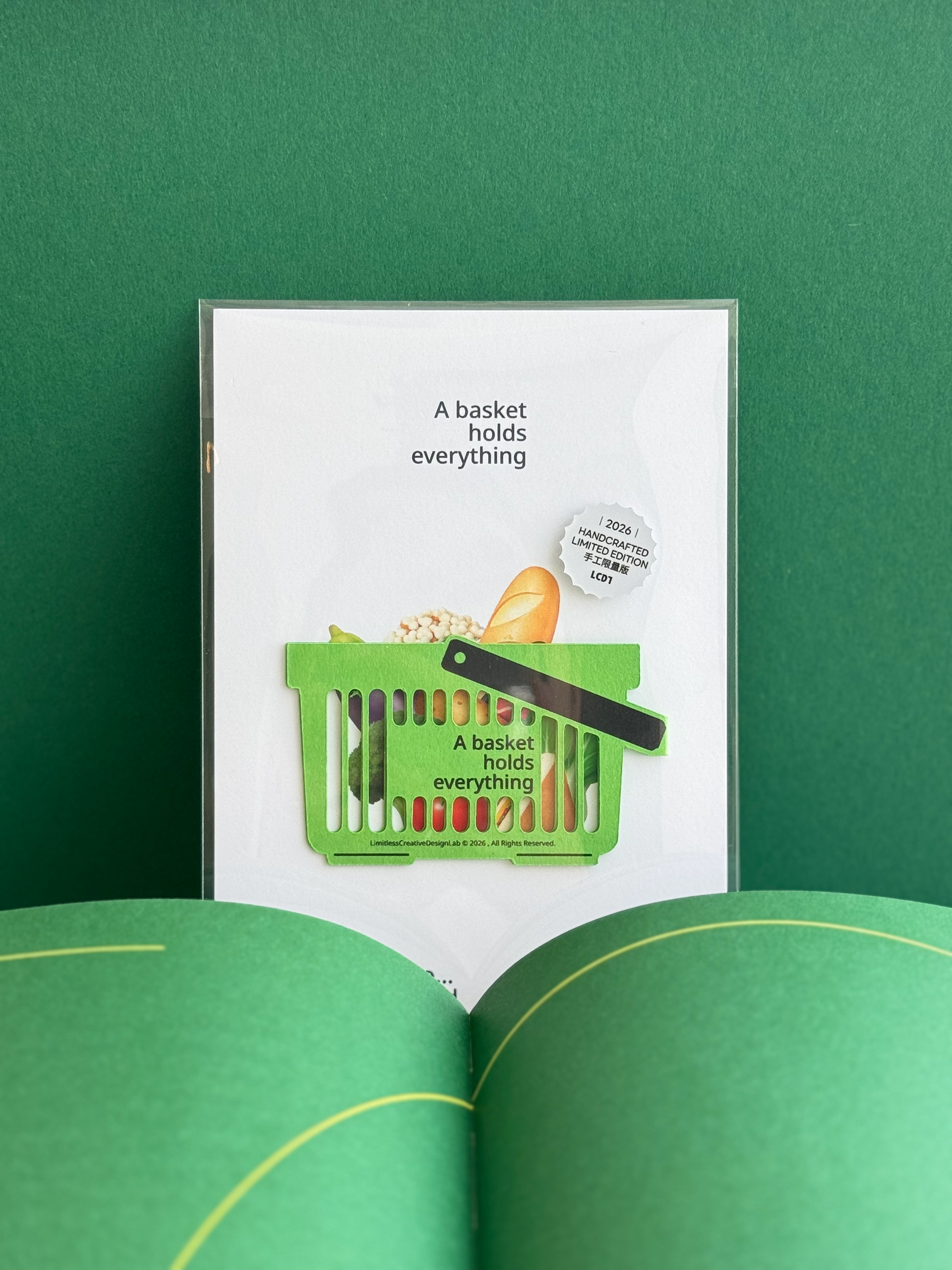 Green shopping-basket bookmark on a white backing card, displayed against a green background and open book.